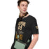 A man wearing a black graphic t-shirt with a Bible verse print, styled with a brown patterned neck scarf and a sage green crossbody bag, set against a plain white background.